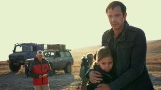 A man is hugging his young daughter. In the background you can see two men, one holding a camera. There are also two large vehicles. They're outside on a deserted, barren landscape.