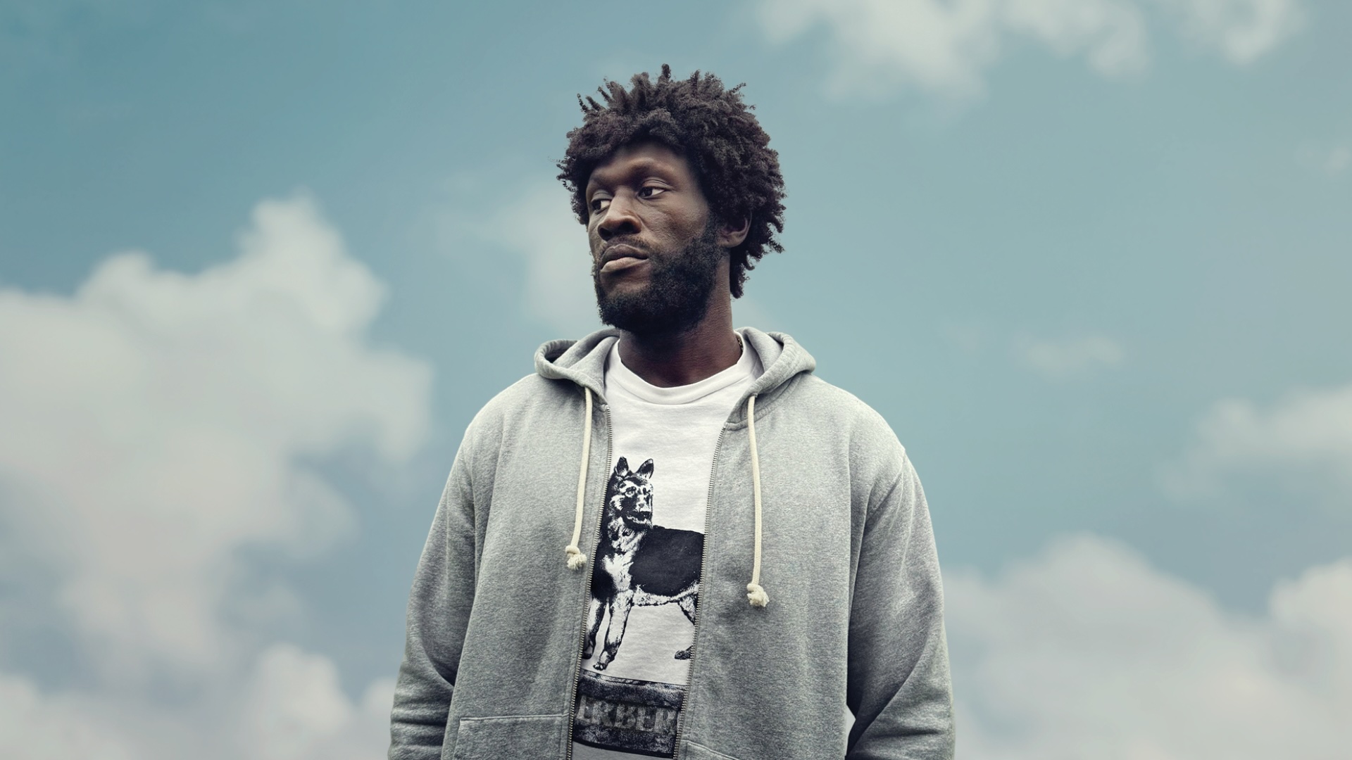 Stormzy standing with a cloudy sky behind him
