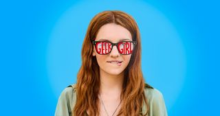 Harriet Carey biting her lip and standing against a blue background wearing glasses with the title 'Geek Girl' written across them in red.