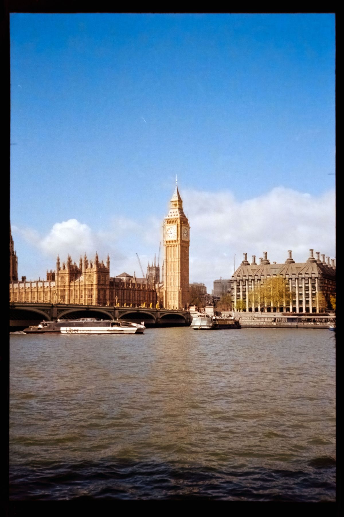 Cropped scan of the Houses of Parliament and Elizabeth Tower across the River Thames under a bright blue sky.
