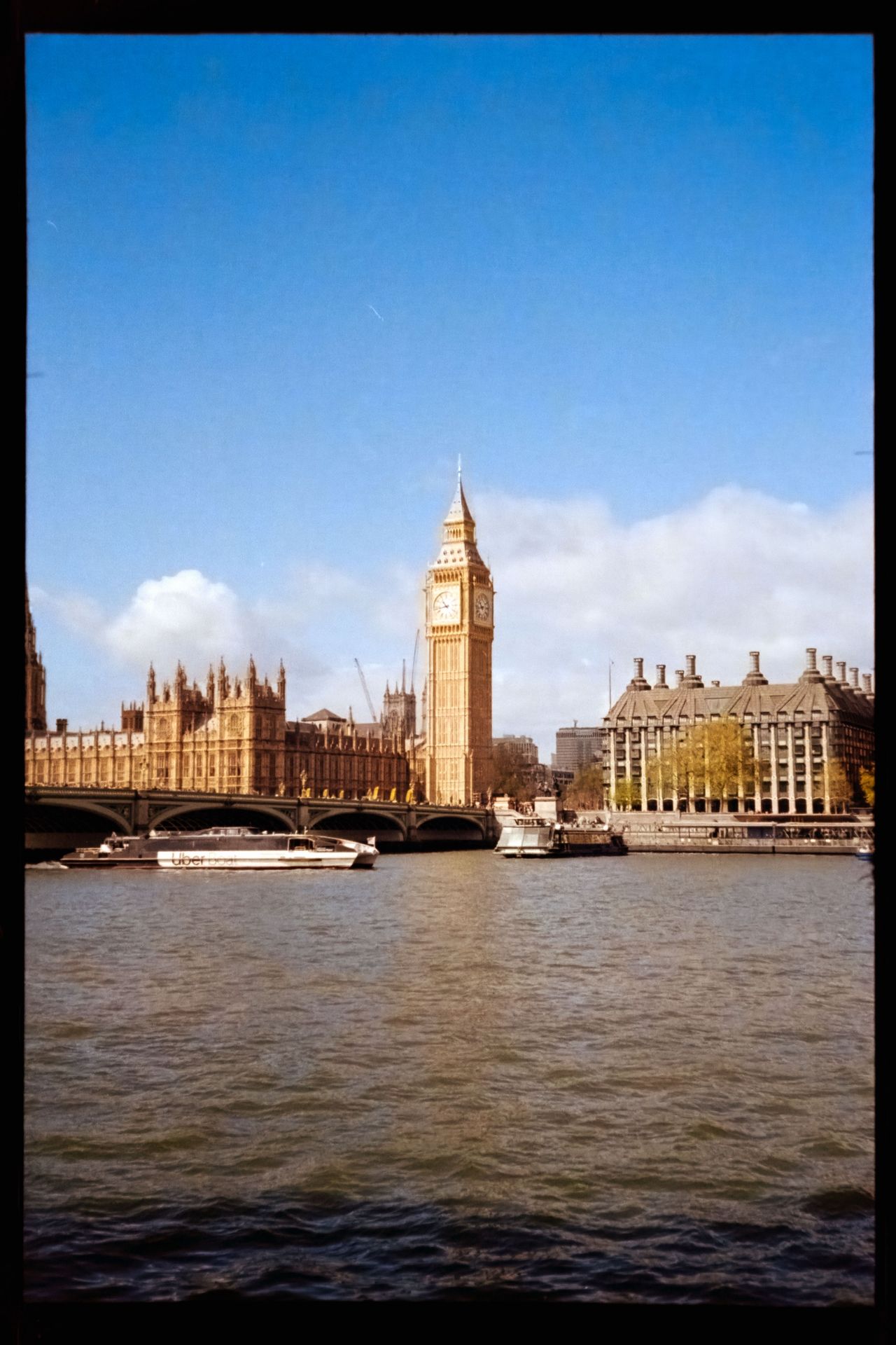 Cropped scan of the Houses of Parliament and Elizabeth Tower across the River Thames under a bright blue sky.
