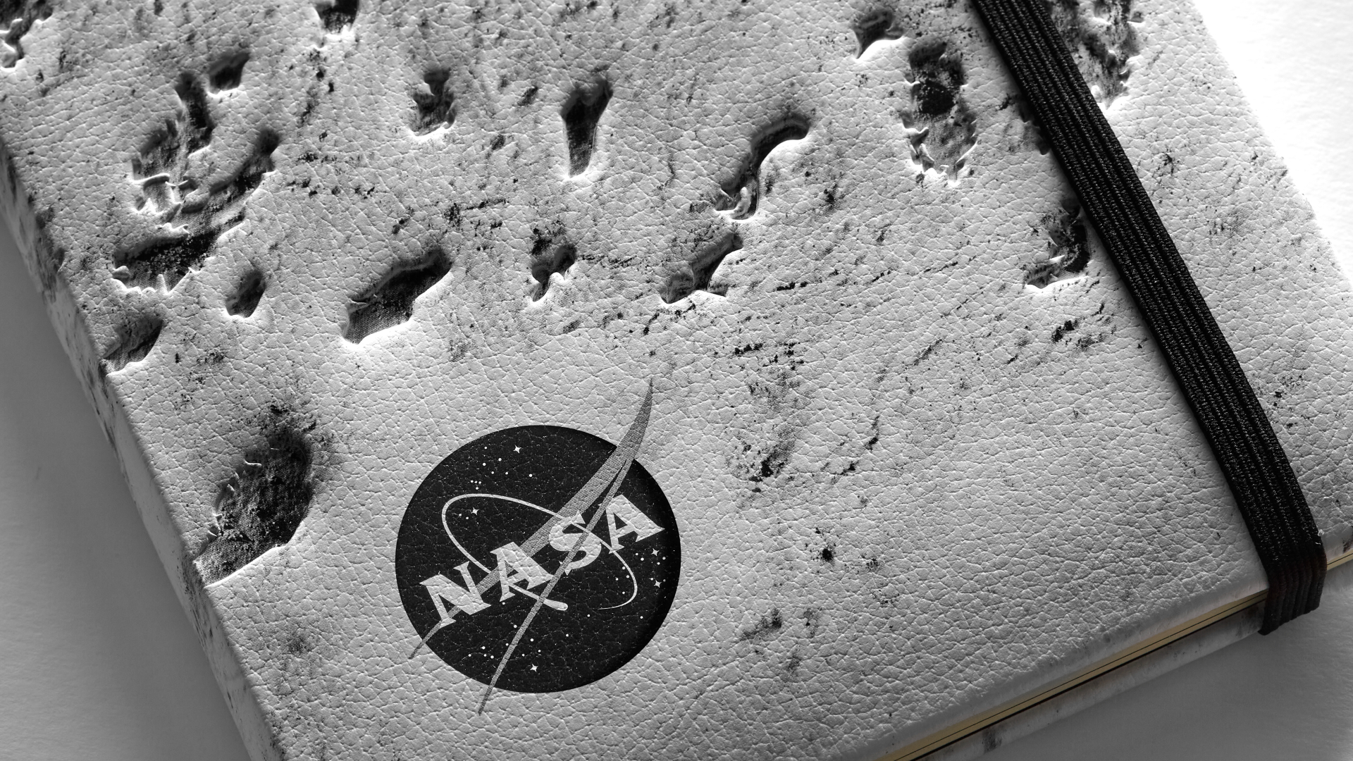 Close up a NASA-inspired Limited Edition Notebook from Moleskine. The cover is a close up of the moon's surface. The notebook has a black elastic closure. A black and white version of the NASA logo is in the bottom left corner.