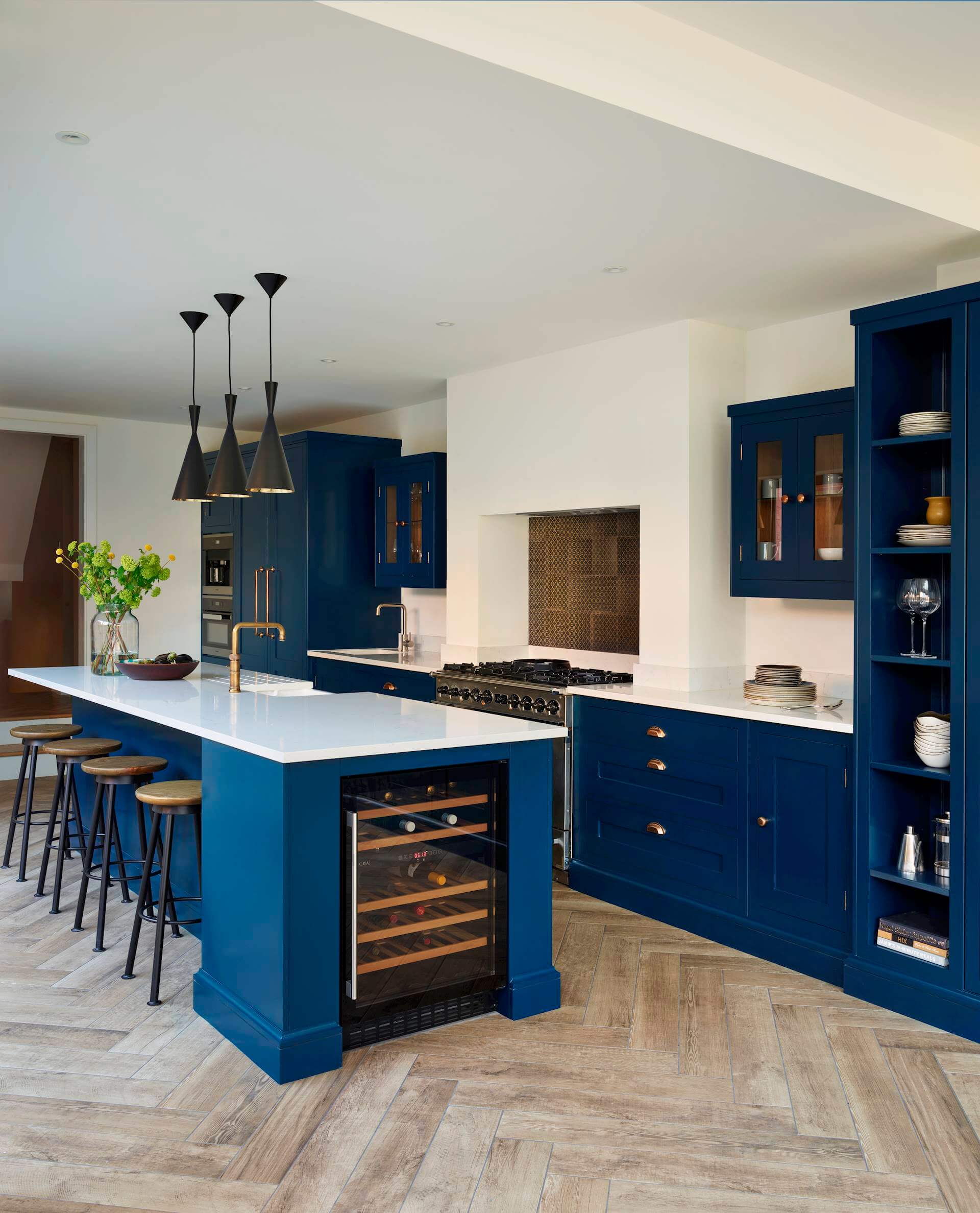 A kitchen by Harvey Jones with blue shaker-style kitchen cabinets and island, wood effect Herringbone flooring, and trio of black ceiling lights over island with four wood and meta bar stools