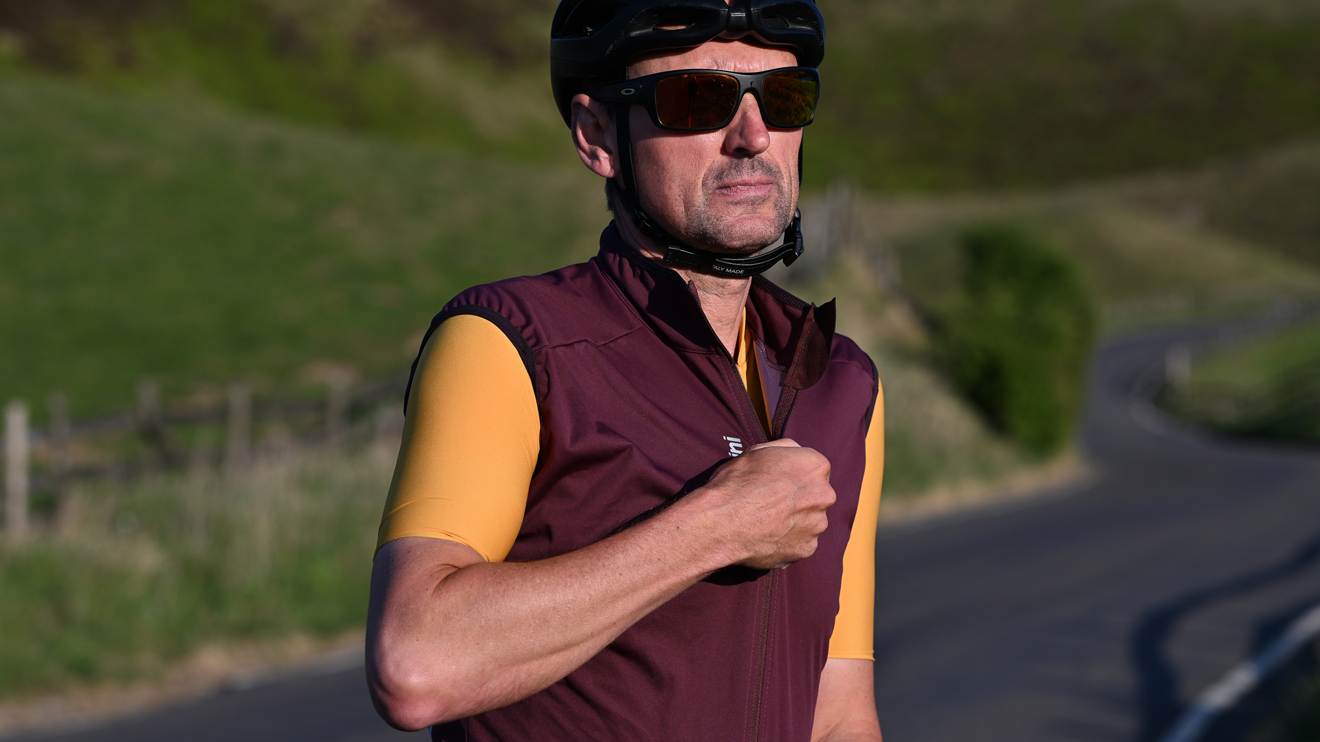 Man wearing a black helmet, sunglasses and a yellow jersey zipping up a purple gilet