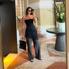 Woman takes a mirror selfie wearing a black asymmetric fringe tube top, brown pants, flip flops and The Row's Barn Tote. 