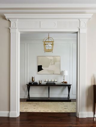 Entry foyer in classical style mouldings with neutral colors and wall paneling wtih modern black console and modern art above brass lantern style lightfitting and pale rug on dark wood flooring