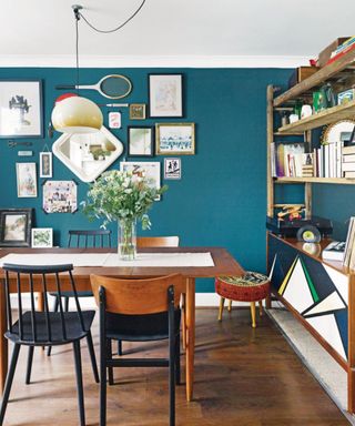 A boho dining room with a teal wall with a gallery wall of prints and sports bats, a hanging pendant light, a warm wooden dining table with four dark blue and wooden chairs either side, and a colorful bookshelf
