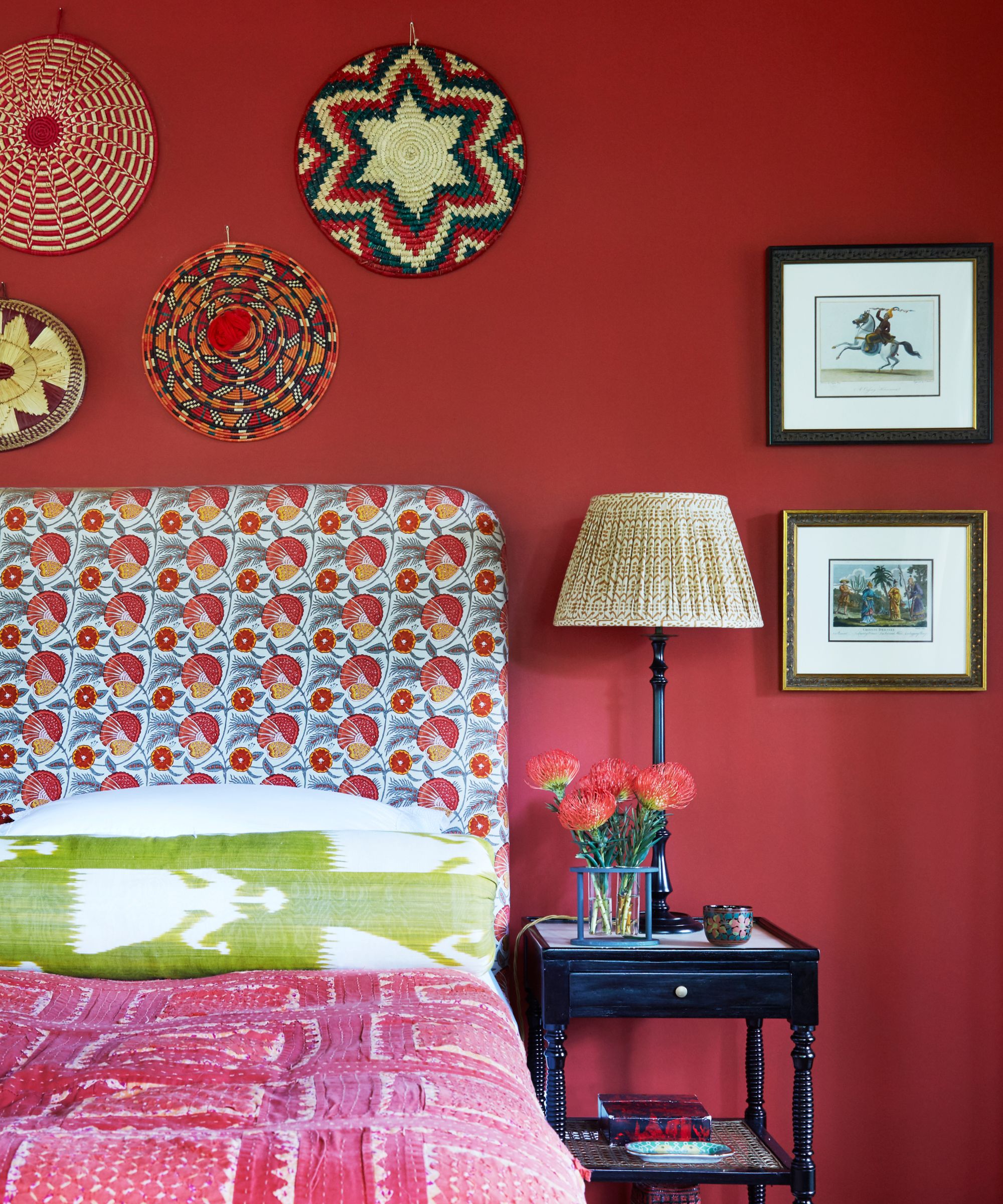 Bedroom with red wall, patterned headboard and pink, white and green bedding. To the right of the bed is a black bedside table with large red flowers, black lamp with white lampshade, and two framed artworks. Above the bed are decorative wall hangings.