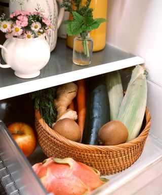 A fridge with two shelves - one with white vases with flowers on and with a woven basket with fresh produce in it and around it