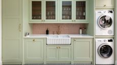 A kitchen with sage green cabinetry, a pink tiled backsplash, and a built-in washer and dryer stacked on top of each other