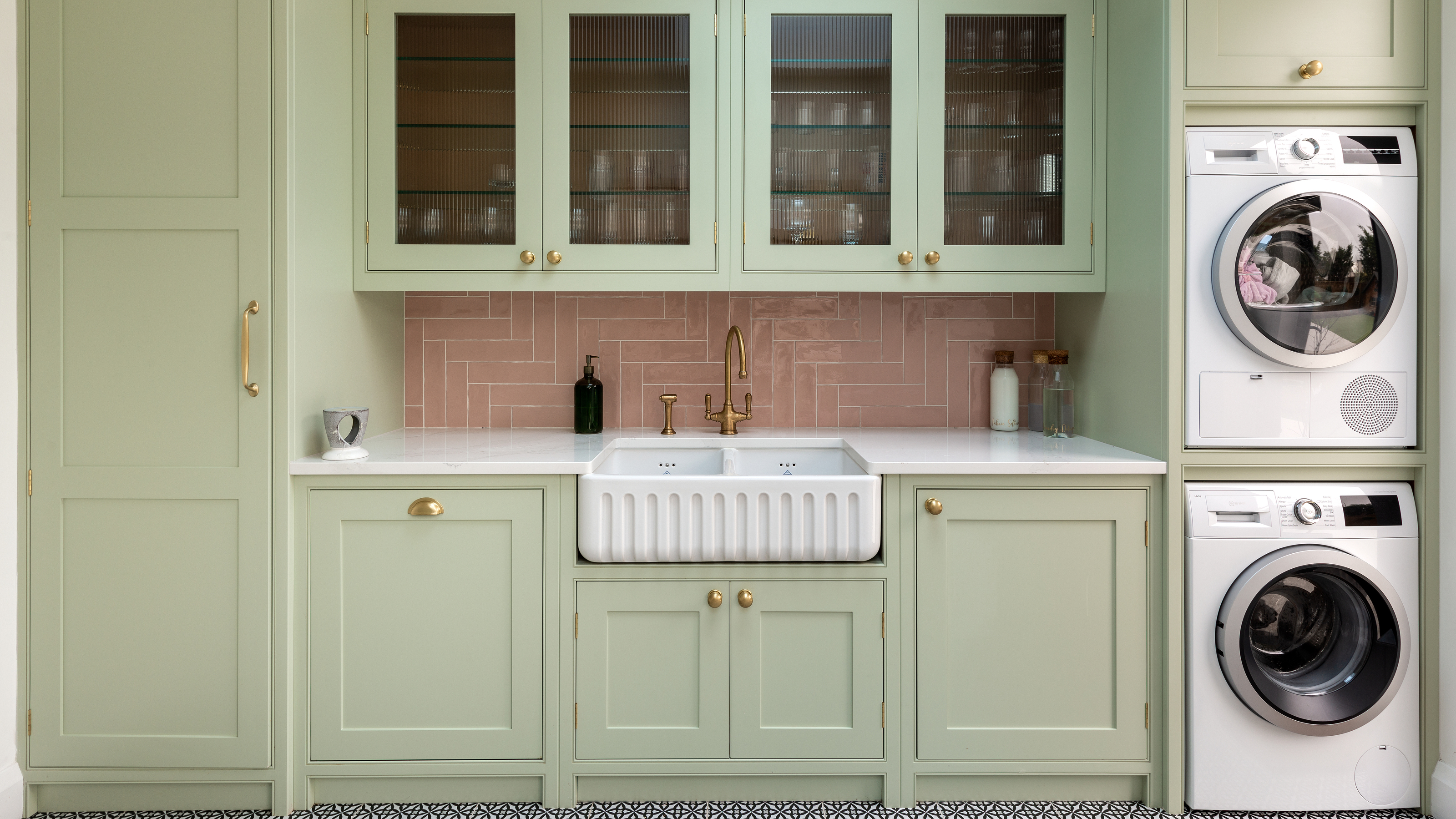 A kitchen with sage green cabinetry, a pink tiled backsplash, and a built-in washer and dryer stacked on top of each other