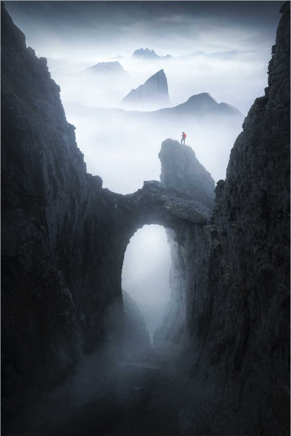A lone figure stands atop a rock formation which is emerging from mist, and there's an arch in the rock below them, seen from an aerial perspective