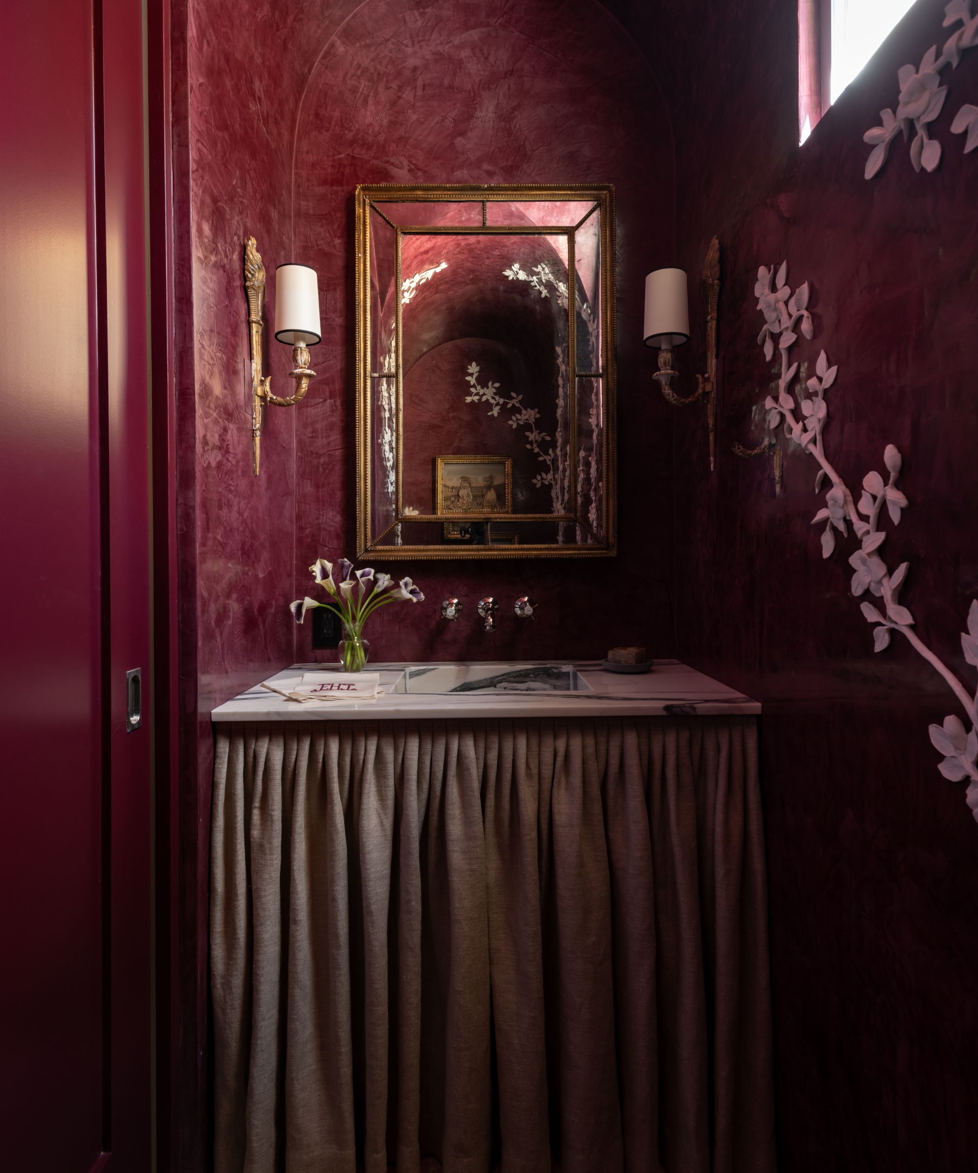 A small powder room with a dark burgundy color scheme. A glossy door and Venetian plaster walls, with white hand-painted flowers on one wall. A sink with a curtain beneath it, a brass mirror, and two decorative wall sconces.