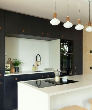 A black kitchen with four pendant lights, a white kitchen island with a silver coffee maker, and black cabinets behind it with a white countertop with a gold sink and plants and decor