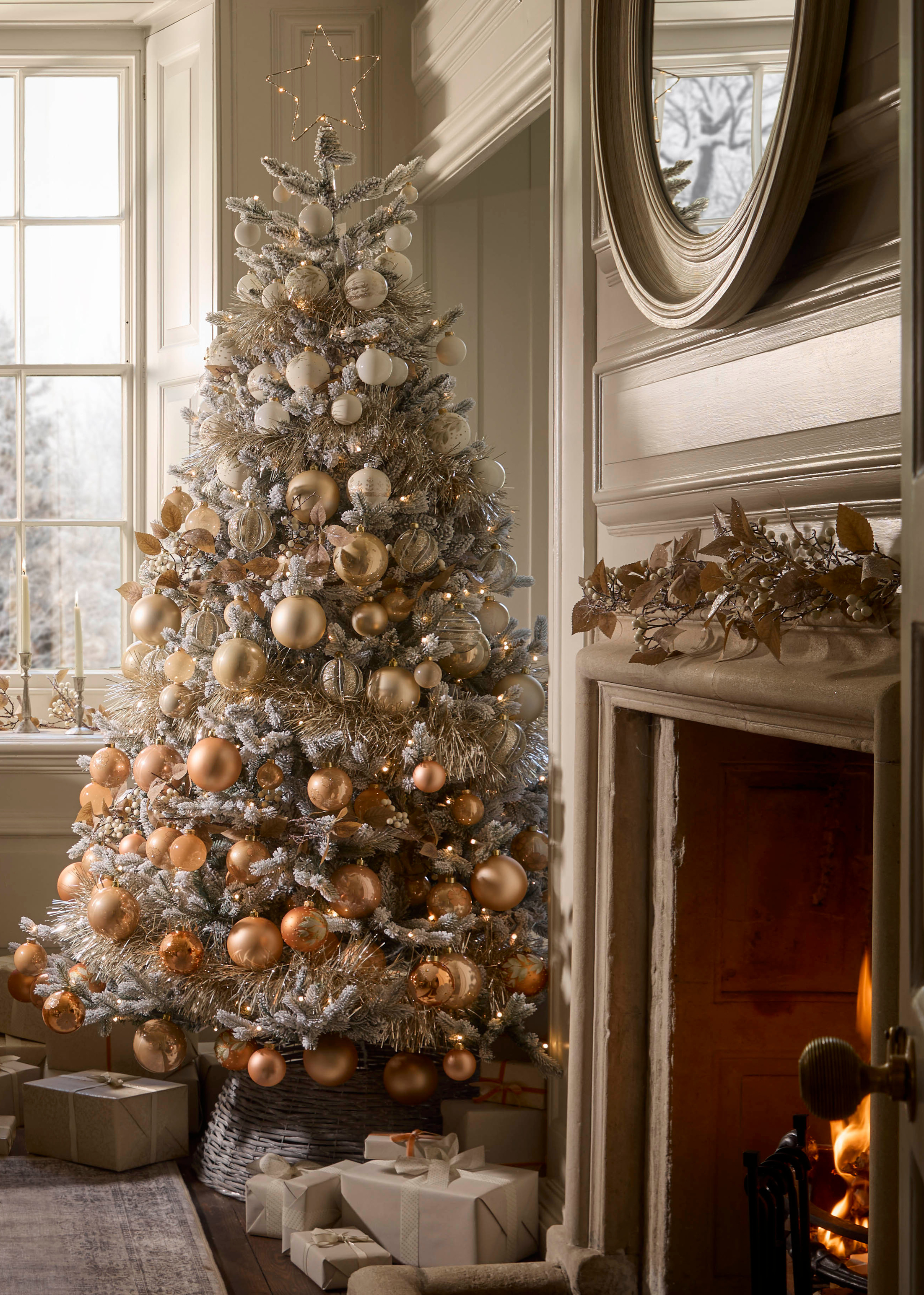 A flocked Christmas tree with neutral baubles by a fireplace and a bundle of wrapped presents