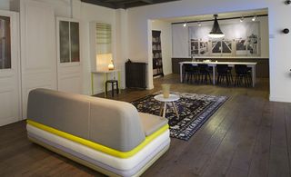 Hôtel Droog pop up, Paris, France. An open plan lounge and dining area with a striped sofa, a round coffee table, a patterned rug, a desk, a heater, a book shelf and a long white dining table with black chairs below are large wall painting.