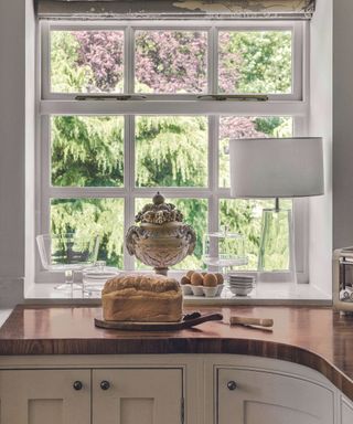 A wood kitchen worktop and curved units, a fresh loaf of bread and fresh eggs and a window out into the garden with a roman blind.