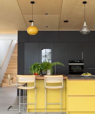 A mid-century modern kitchen with two hanging globe lights - one yellow one glass - a black wall, a bright yellow kitchen island with plants on, and two light wooden rattan bar chairs in front of it