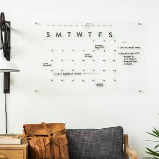 A clear acrylic wall calendar with the days of the week and a grid printed on. Black pen handwriting across it