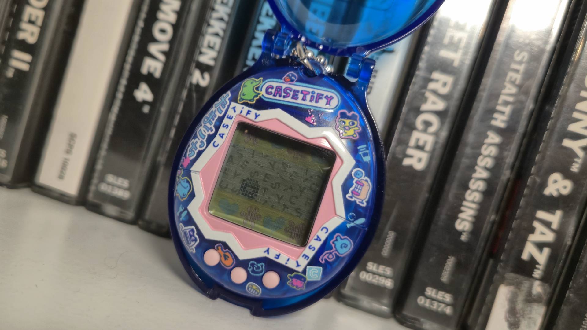 Photo of the Casetify Tamagotchi sitting in front of a row of PlayStation games.