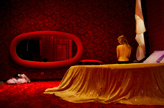 Greek sex worker topless with her back turned, sit on a bed in a plush red room.