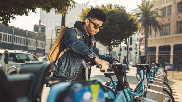 A man getting a bike from a bike rack, wearing a pair of the best sunglasses for men, a denim jacket and a backpack