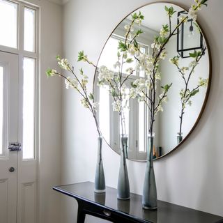 White hallway with thin dark brown hallway table with three grey vases with white flowers in it in front of round mirror