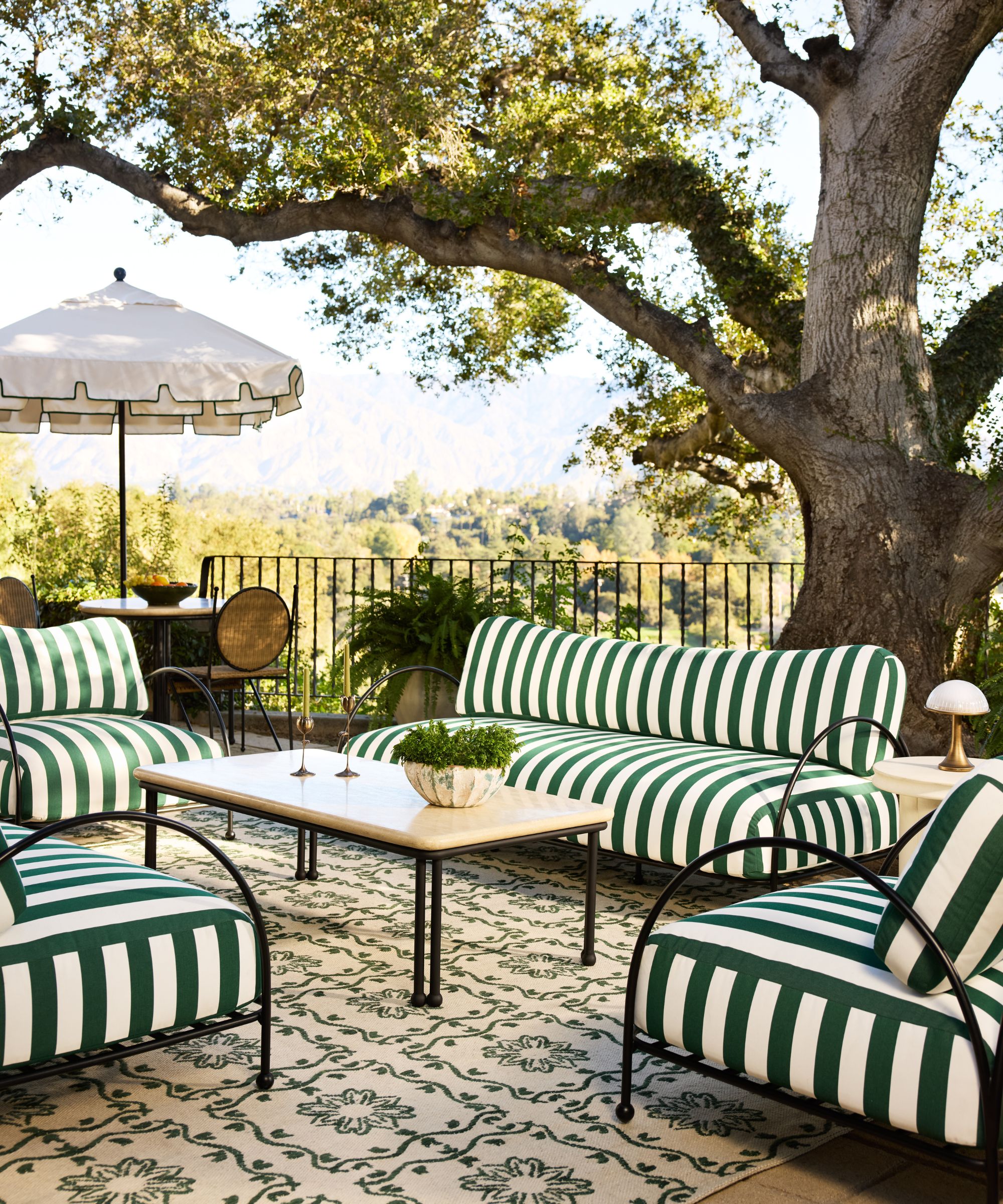 a european style terrace with a large tree, and an arrangement of green striped wrought iron furniture on a patterned green floral rug