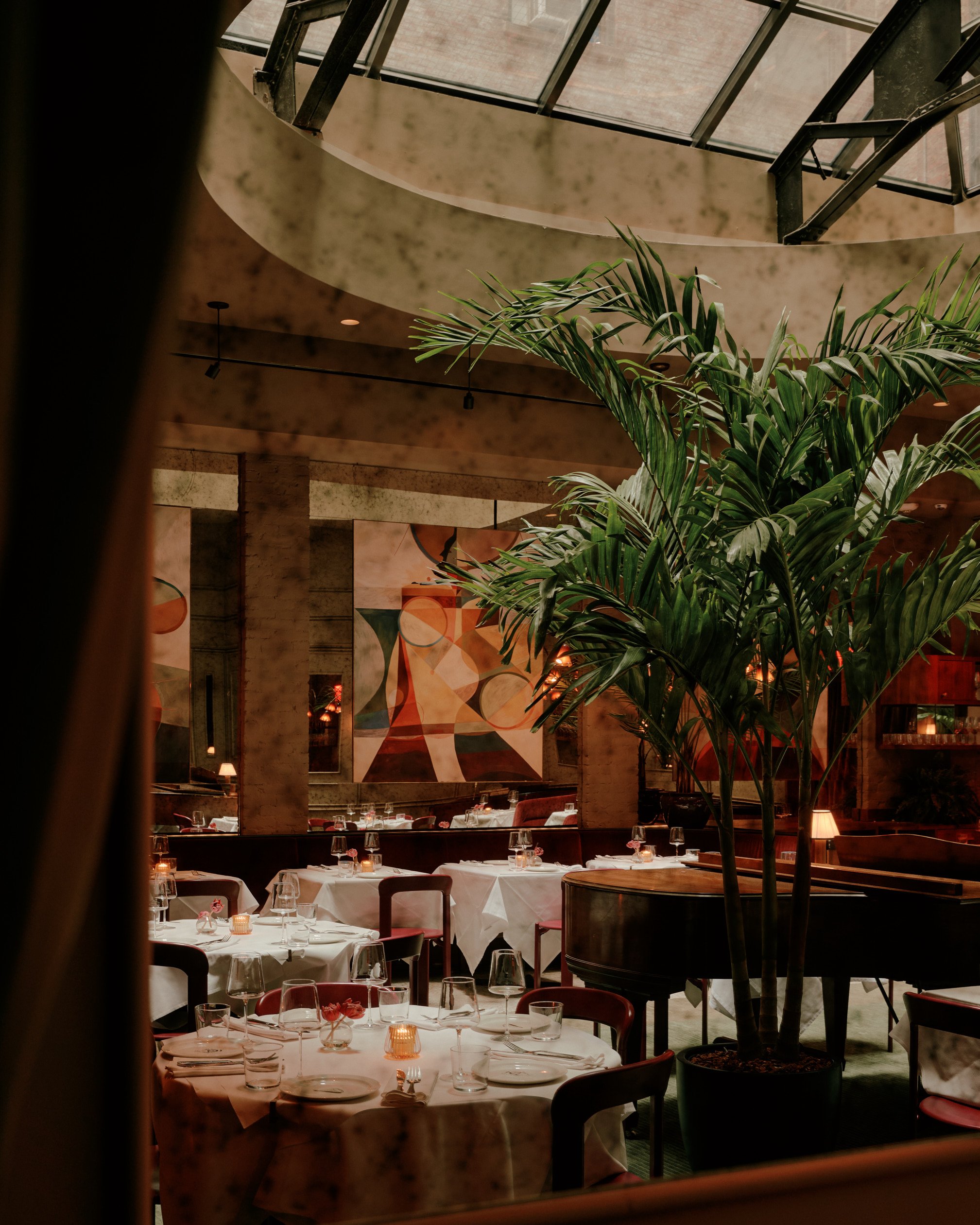 A dimly lit, imaginatively decorated cocktail bar features murals, paintings, Art Deco-inspired lighting, and sprawling plants in a warmly suffused atmosphere.