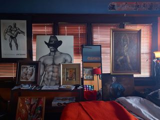Bedroom interior, corner of bed with white sheets and red cover, black framed windows with red blinds, black table under window, books on table, corner light, male erotic framed art on the walls and table, hung in front of the windows and propped up against walls and furniture on the floor, slim black framed floor mirror, reflecting art, tall red standing lamp and another angle of the room