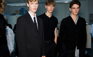 Male models wearing Margaret Howell S/S 2015 collection. The model on the left is wearing a black suit and white shirt. The model next to him is wearing a black shirt and black pants, while the model on the right is wearing a black overcoat.