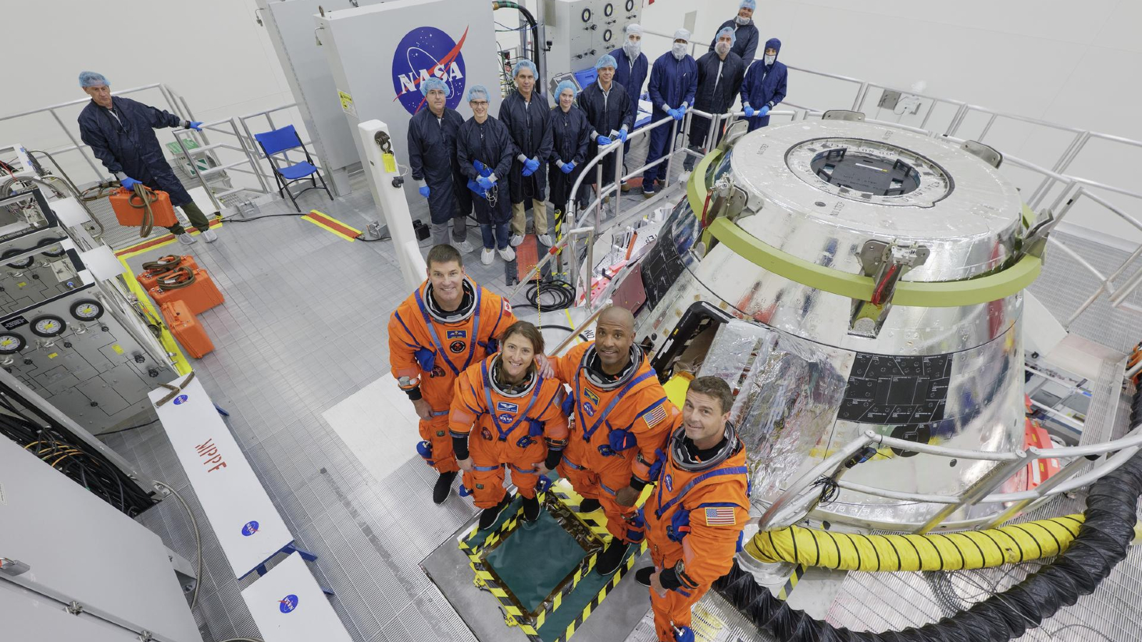 A photo of the Artemis II crew stood in front of the Orion capsule with around a dozen technicians in a NASA clean room