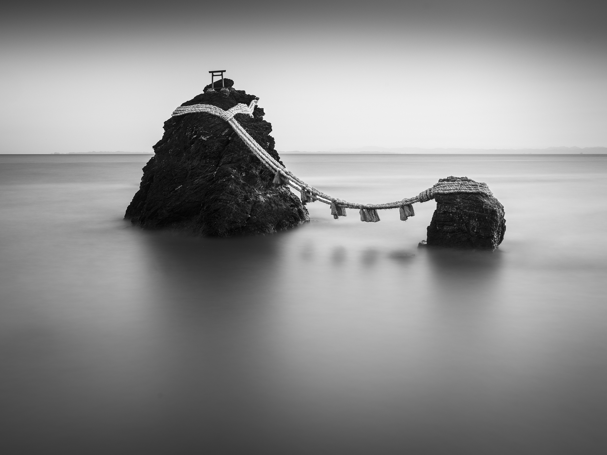 A serene black-and-white image of a rocky islet with a traditional torii gate, connected by a straw rope and surrounded by calm waters