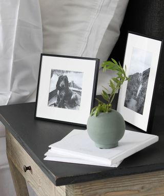 A black nightstand with two photo frames and a plant on it, next to white bedding