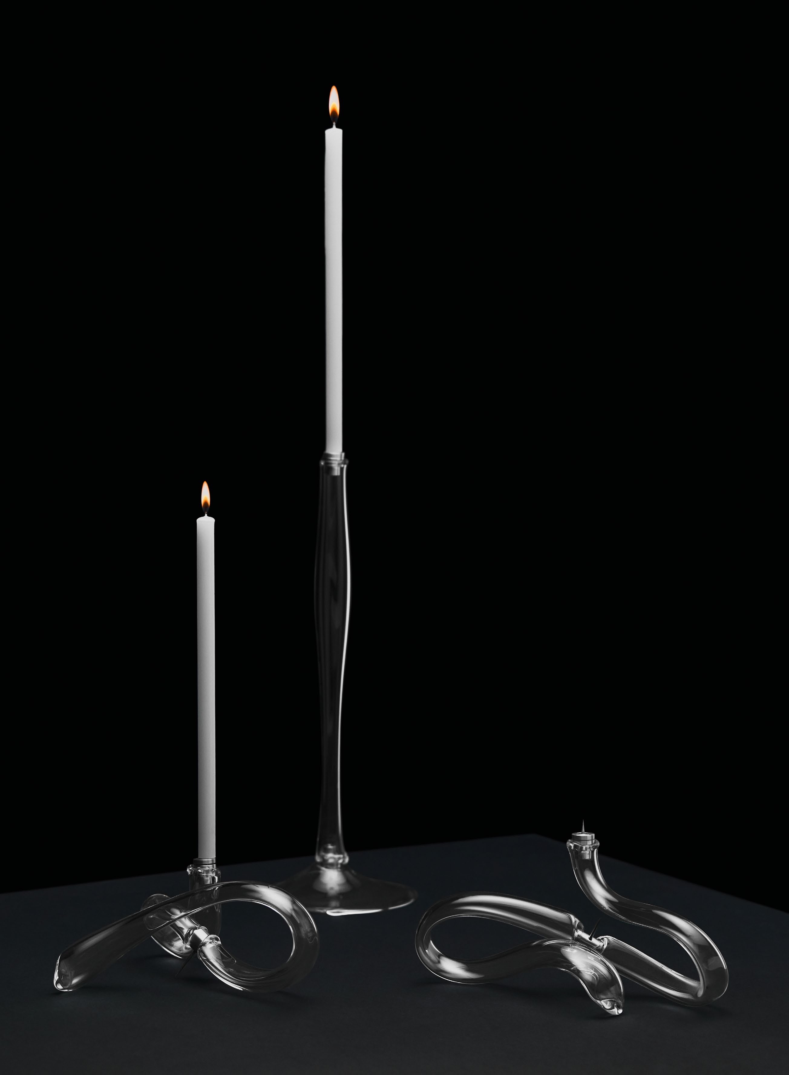 A contemporary glass candelabra series designed by Miranda Keyes and Karum Friel, featuring elegant glass candle holders with lit candles against a dark background.