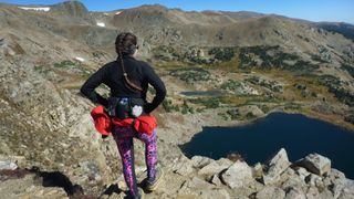 A hiker wearing a waist pack with water bottles looks down at an alpine lake in the Rocky Mountains