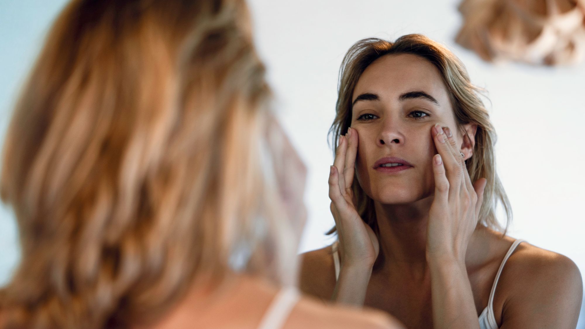 woman looking in the mirror with hands on her cheekbones - unexpected signs of ageing