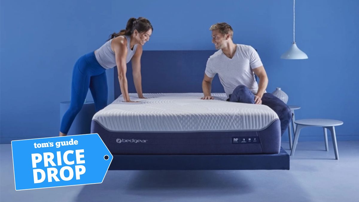 A couple sitting on a Bedgear mattress with a woman standing on the left and a man sitting on the right, a Tom&#039;s Guide Price Drop tag in the lower left corner