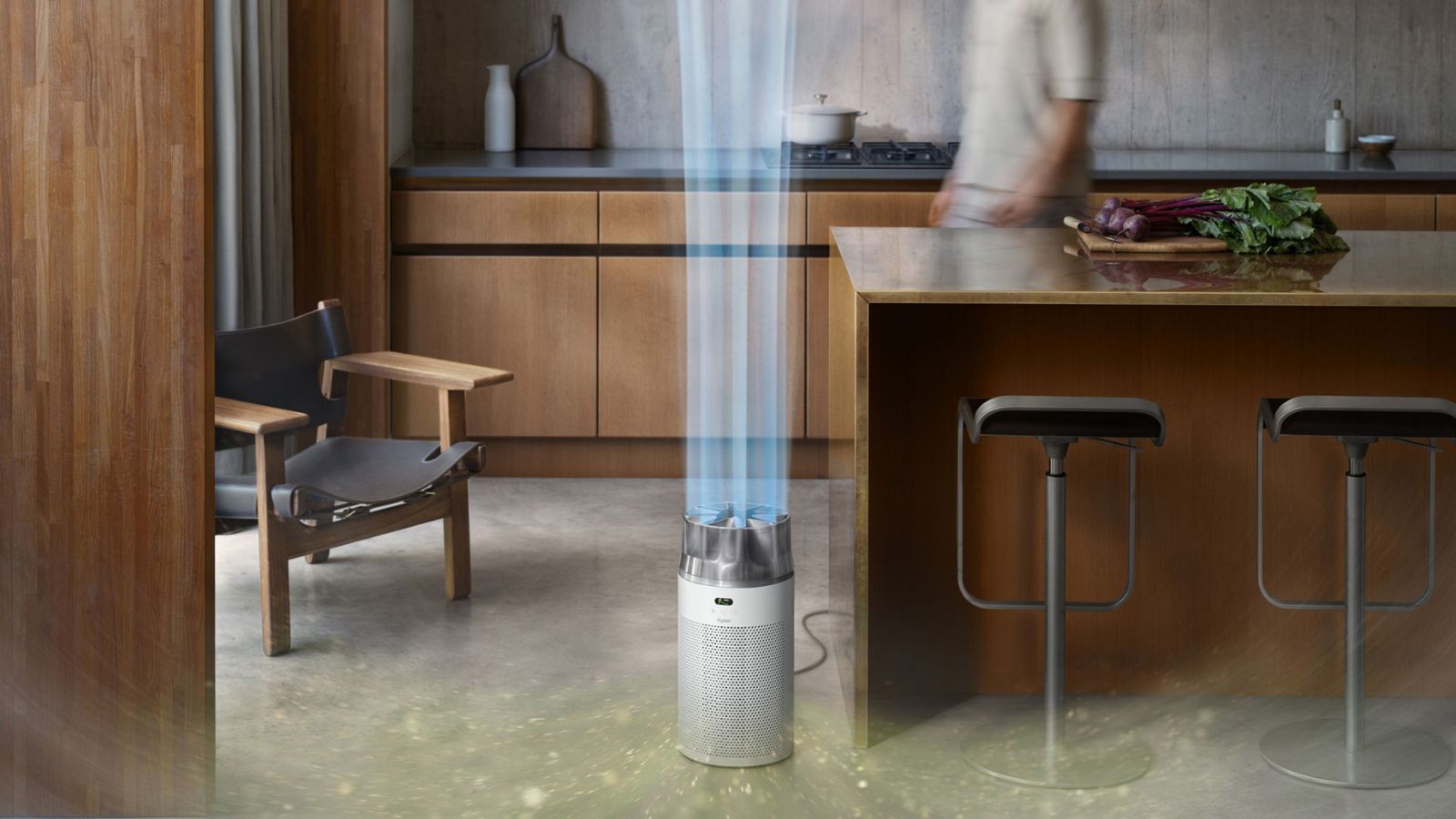 The white Dyson Hushjet Purifier Compact seen in a modern kitchen with wooden cabinetry, a wooden island with metal stools, a Scandi-style dark grey and wood chair, and dark grey countertops with a light beige panelled backsplash behind.