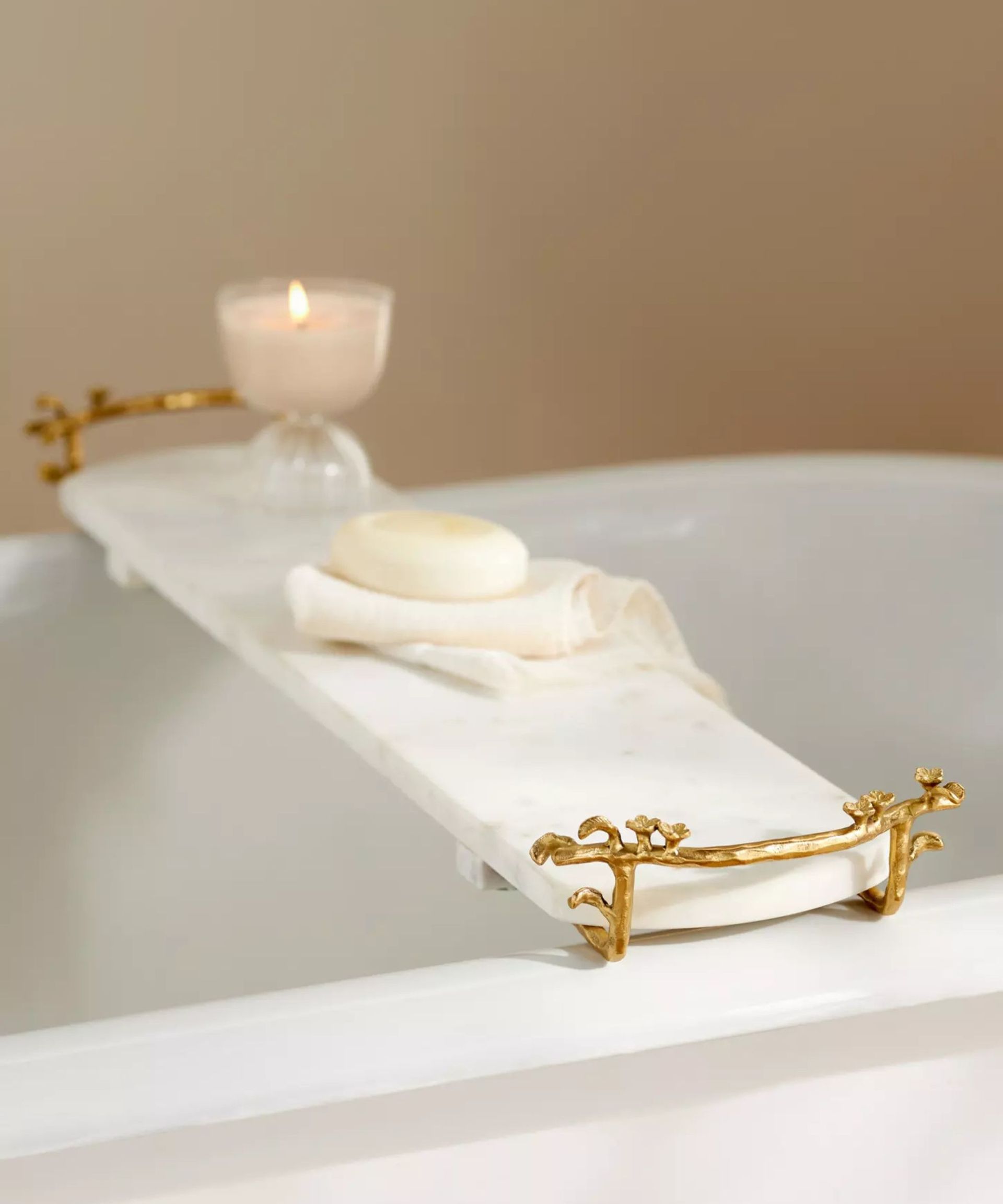 A white marble bath caddy with very light gray veins. It has gold ornate handles either side and is placed over a roll-top white bath, with a lit candle and soap placed on a towel