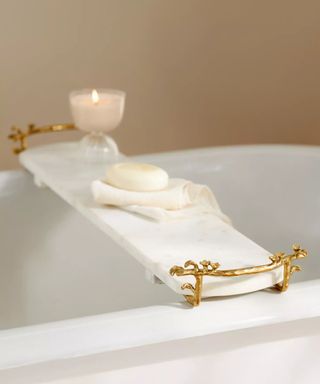 A white marble bath caddy with very light gray veins. It has gold ornate handles either side and is placed over a roll-top white bath, with a lit candle and soap placed on a towel