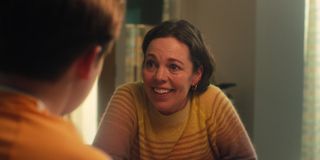 Olivia Colman as Sarah Nelson in Heartstopper, wearing a brown and beige striped jumper. She is seated at a table and smiling at her son Nick (Kit Connor) - we can see the back of his head out of focus in the foreground