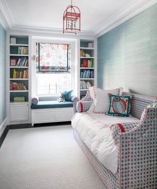 A kids bedroom with blue grasscloth wallpapered walls, blue and red patterned daybed, and a window seat