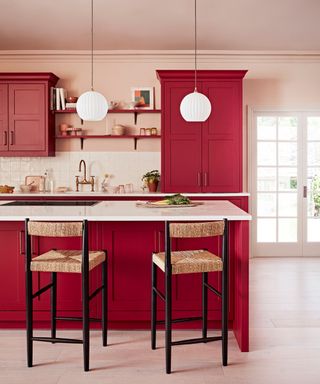 A kitchen with deep red cabinets and shelves, two white circular pendant lights, a kitchen island with a hob, a white counter, and two rattan chairs in front of it