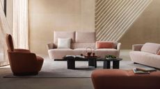 open plan living room with sandstone walls, light shining in, sheer curtains, a light pink sofa from King Living with the backrest flipped up, two black coffee tables, another pink sofa, an orange armchair and ottoman and a gray rug on the floor