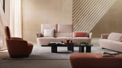 open plan living room with sandstone walls, light shining in, sheer curtains, a light pink sofa from King Living with the backrest flipped up, two black coffee tables, another pink sofa, an orange armchair and ottoman and a gray rug on the floor