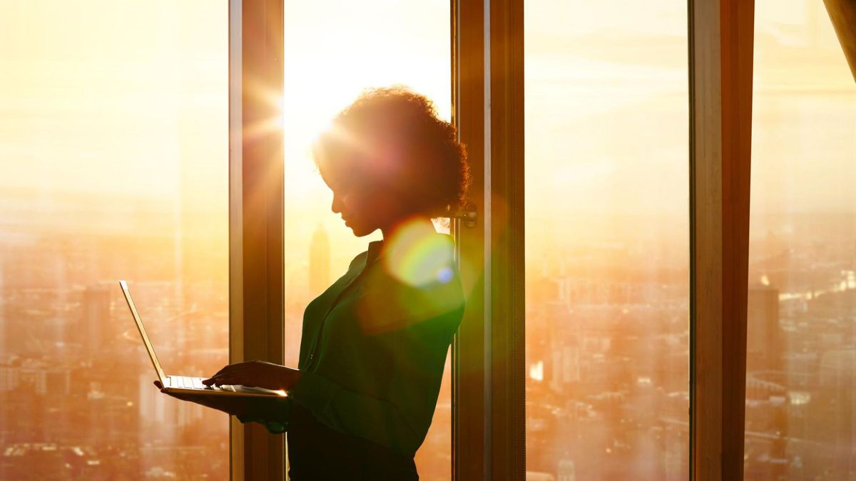 Woman holding laptop and standing at office window with a cityscape beyond