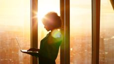 Woman holding laptop and standing at office window with a cityscape beyond