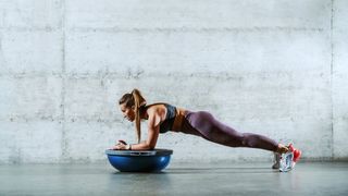 Woman performing a forearm plank on the Bosu balance trainer against a grey backdrop