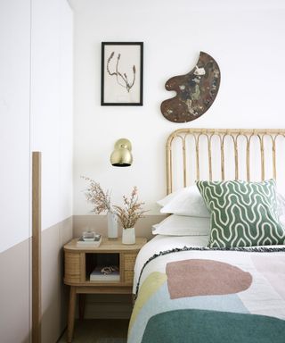 Bright white bedroom with lower half the wall painted in a light brown, bamboo style bed-frame, white bed linen with green patterned cushions, pastel throw with geometric shapes, wooden bedside table with small vases of flowers, metallic wall light above, framed artwork and painters palette mounted above bed,